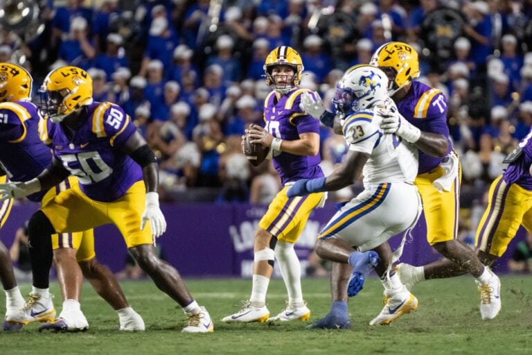 LSU players in purple and gold uniforms protect their quarterback as he prepares to throw a pass.
