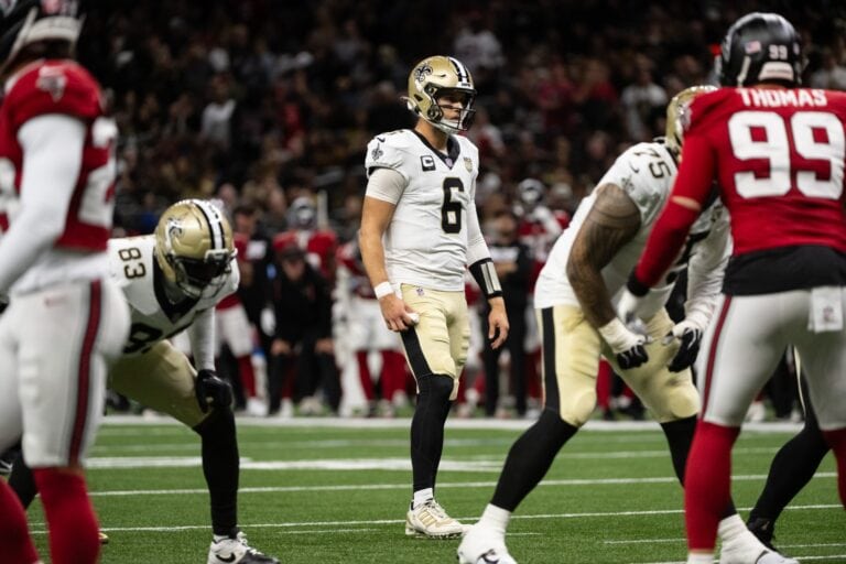 A New Orleans Saints quarterback stands on the field, surrounded by teammates and opposing players in an NFL stadium.