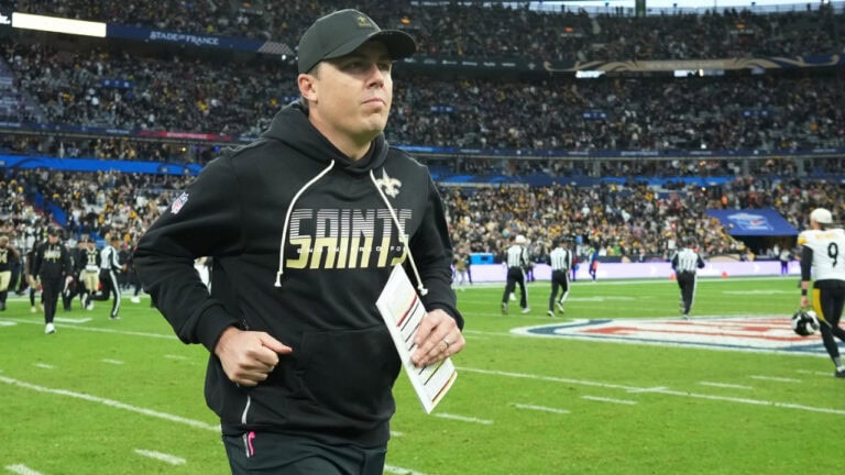 A New Orleans Saints coach in a black hoodie runs across the field holding play sheets while fans watch the game.