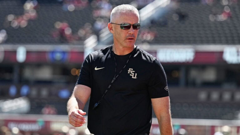 College football coach Mike Norvell stands on a stadium field wearing sunglasses and a black Florida State shirt.