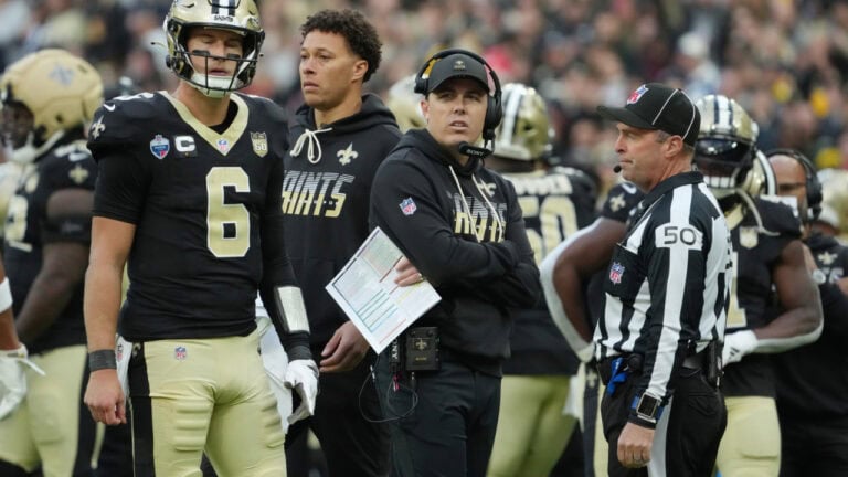 New Orleans Saints players and coach Kellen Moore stand on the field with an NFL referee during a game.