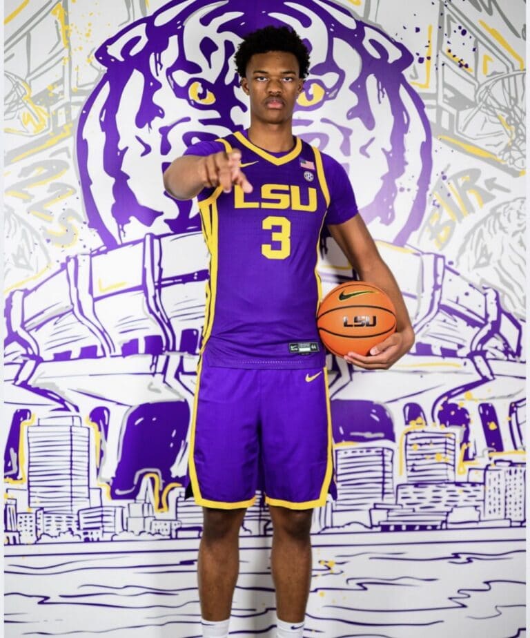 LSU basketball player Xavier Young in his purple and yellow uniform holds a ball and points toward the camera.