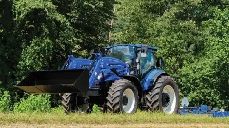 A blue New Holland tractor with a front loader parked in a grassy field in front of trees for winter storage.